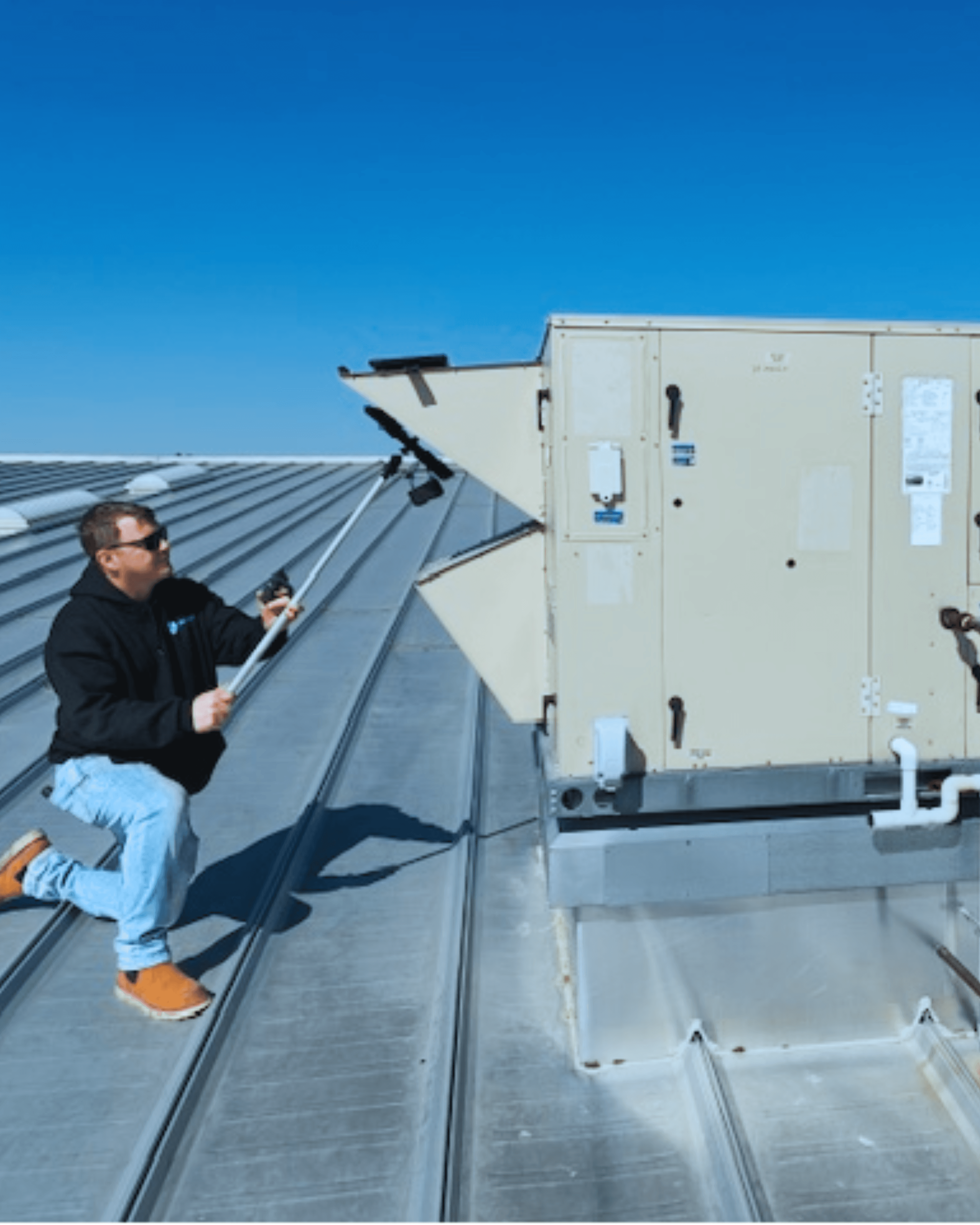 A man kneels on a metal roof, inspecting a large HVAC unit using tools under a clear blue sky.