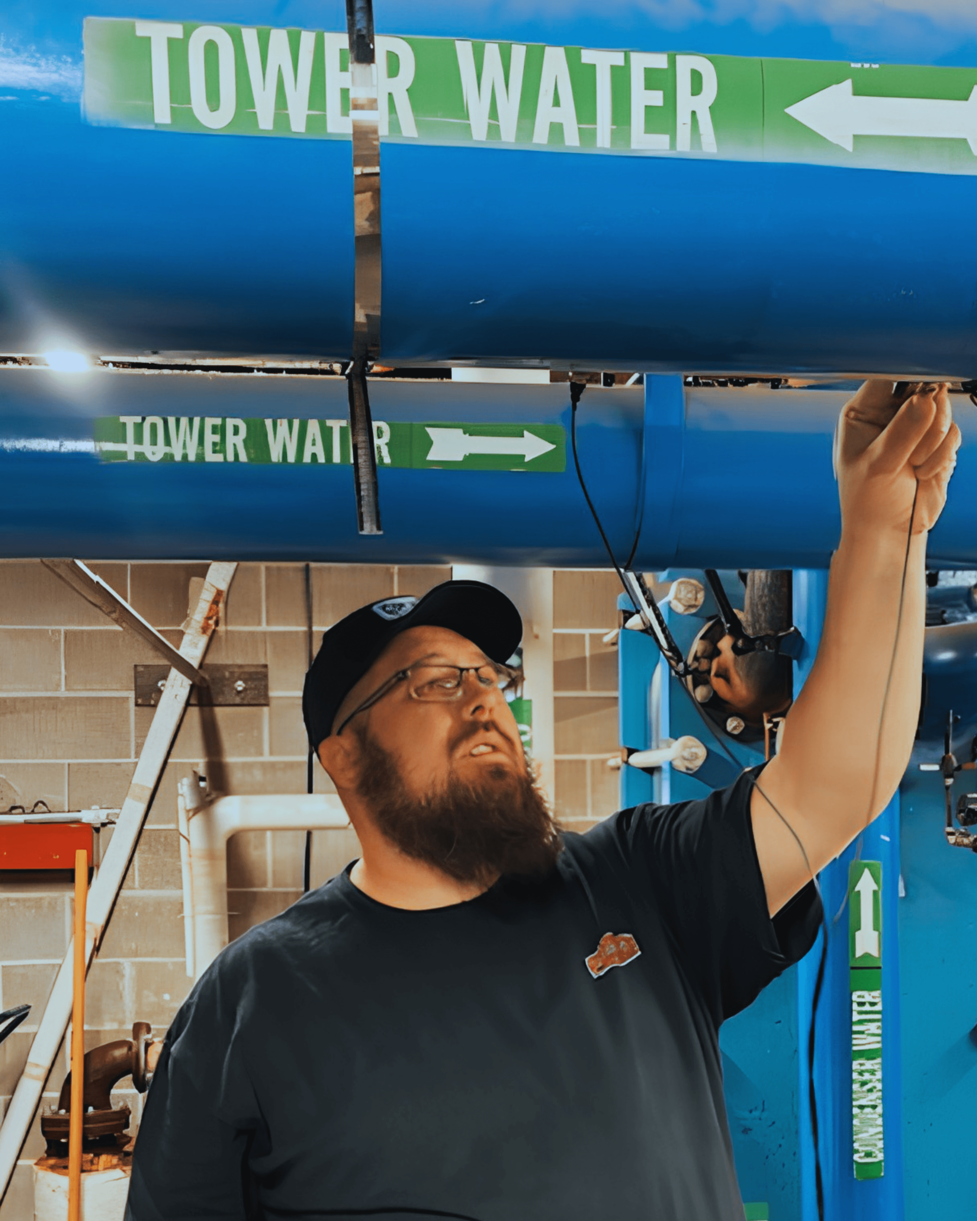 A person in a cap examines a blue pipe labeled "Tower Water" in an industrial setting, pointing at a connected wire.